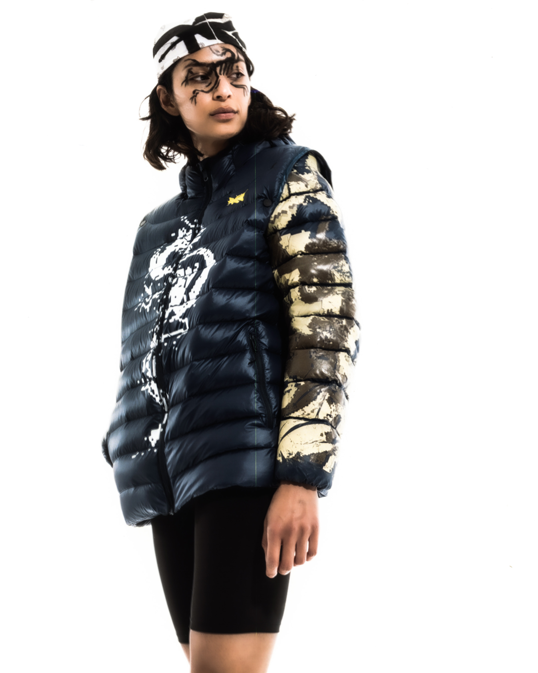 Portrait of a model wearing glack graphic liner, a puffer vest and bandana against a white background